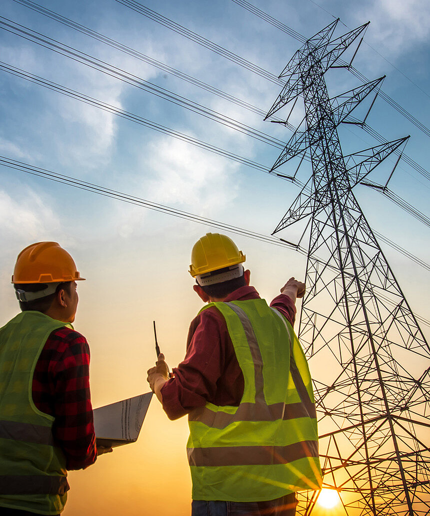Zwei Bauarbeiter stehen vor einem Hochspannungsmast