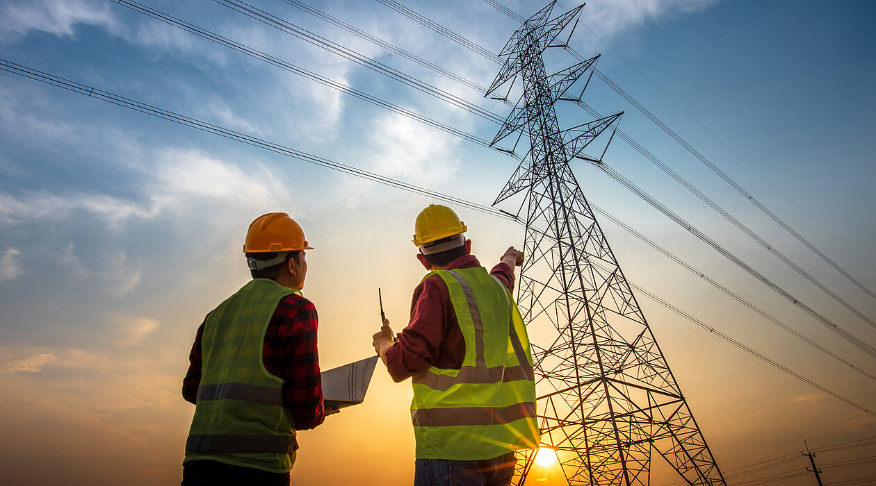 Zwei Bauarbeiter stehen vor einem Hochspannungsmast