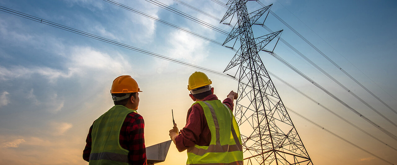 Zwei Bauarbeiter stehen vor einem Hochspannungsmast